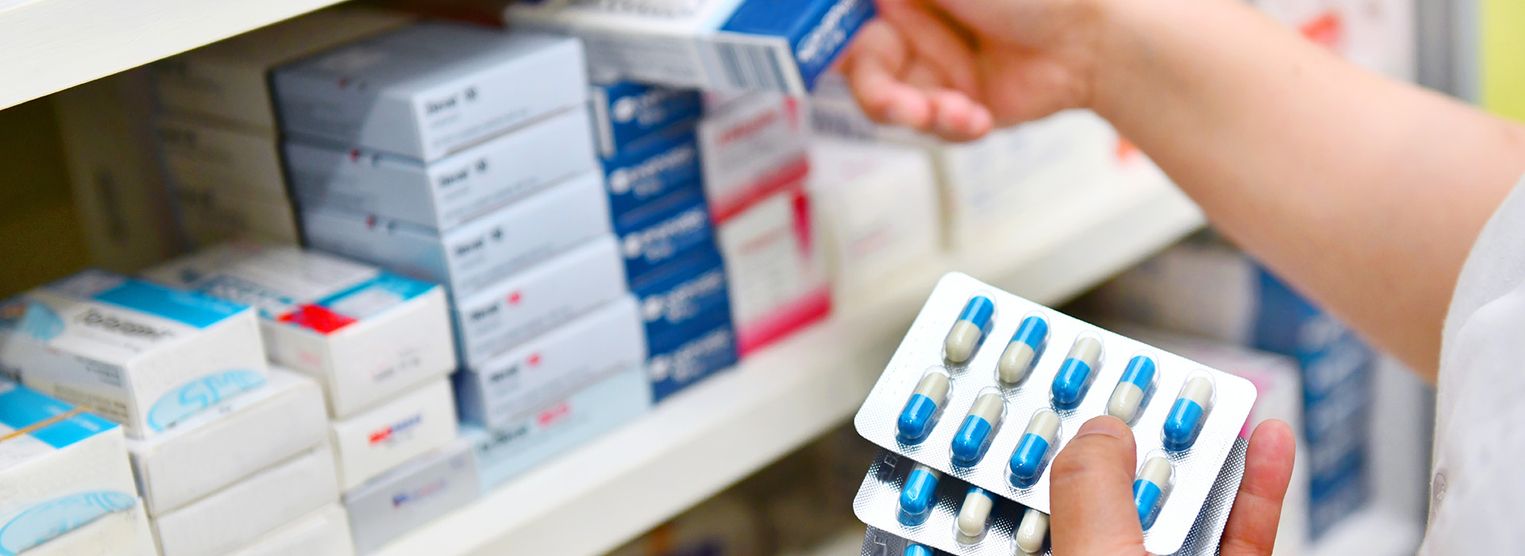 Eine Person hält eine Blisterpackung mit blauen und weißen Kapseln in der Hand, während sie nach einer Medikamentenschachtel im Regal einer Apotheke greift.
